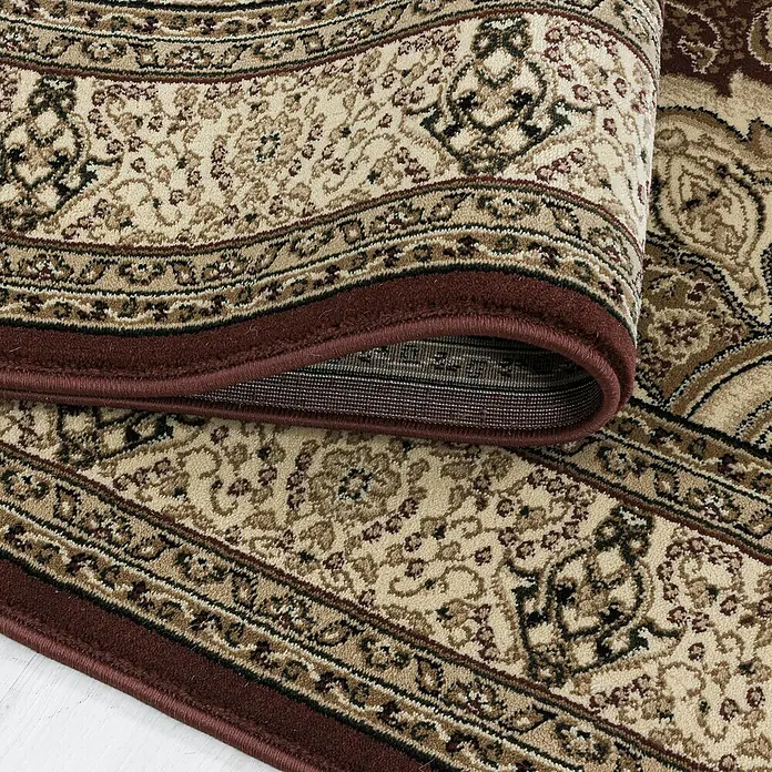 Kurzflorteppich Kashmir 2601Rot, 150 x 80 cm, 100% Polypropylen - Nahaufnahme eines eleganten Teppichs mit floralem Muster und bordeauxrotem Rand, teilweise aufgerollt.