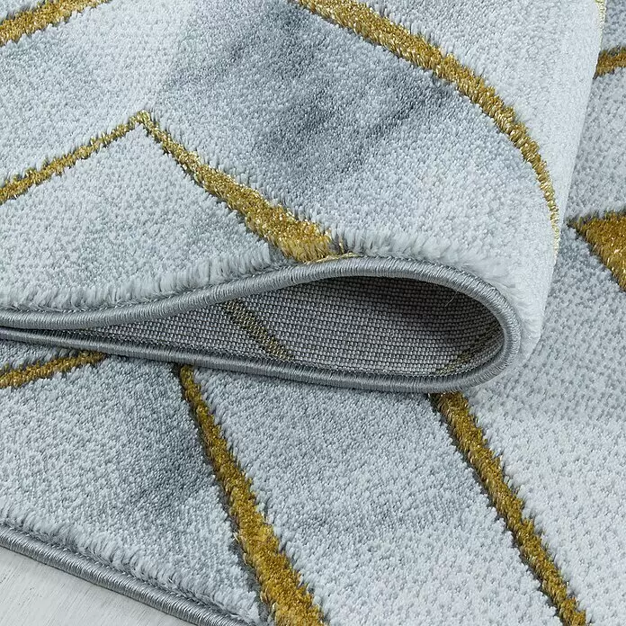 Kurzflorteppich Naxos 3811 - Gold, 200 x 140 cm, 80% Polypropylen, 20% Polyester - Ein plüschiger grau-weißer Teppich mit goldenen Diagonalstreifen, teilweise aufgerollt, zeigt die gewebte Rückseite und den fertigen Rand.