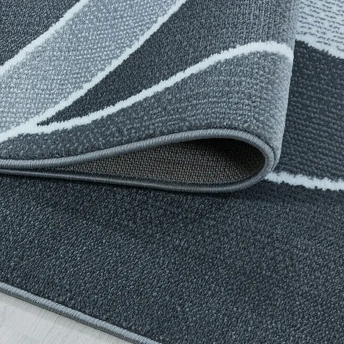 Kurzflorteppich Costa 3523Grau, 290 x 200 cm, 100 % Polypropylen - Nahaufnahme eines modernen grauen Teppichs mit geometrischen Formen, teilweise aufgerollt, um seine Textur und Verarbeitung zu zeigen.
