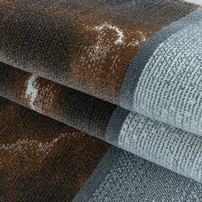 Kurzflorteppich Ottawa 4201 - Kupfer, 200 x 140 cm, 100% Polypropylen - Detailaufnahme eines gefalteten Teppichs mit braunen, grauen und weißen Farbtönen, die ein abstraktes Muster bilden. Die Textur ist deutlich erkennbar.