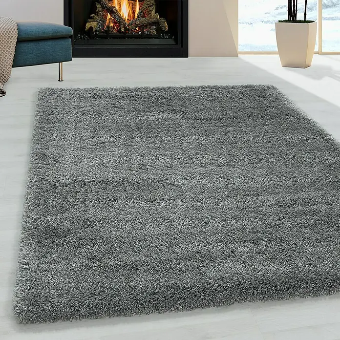 Hochflorteppich Fluffy 3500Light Grey, 370 x 380 cm, 100% Polypropylen - Ein grauer, hochfloriger Teppich liegt in einem Wohnzimmer mit Kamin und Sessel.