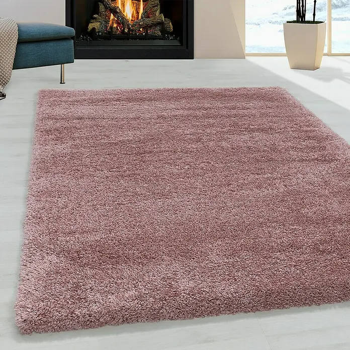 Hochflorteppich Fluffy 3500Rose, 370 x 380 cm, 100 % Polypropylen - Ein quadratischer, zotteliger Teppich in einem warmen Rosaton liegt auf einem hellen Holzboden vor einem Kamin und einem blauen Sessel.