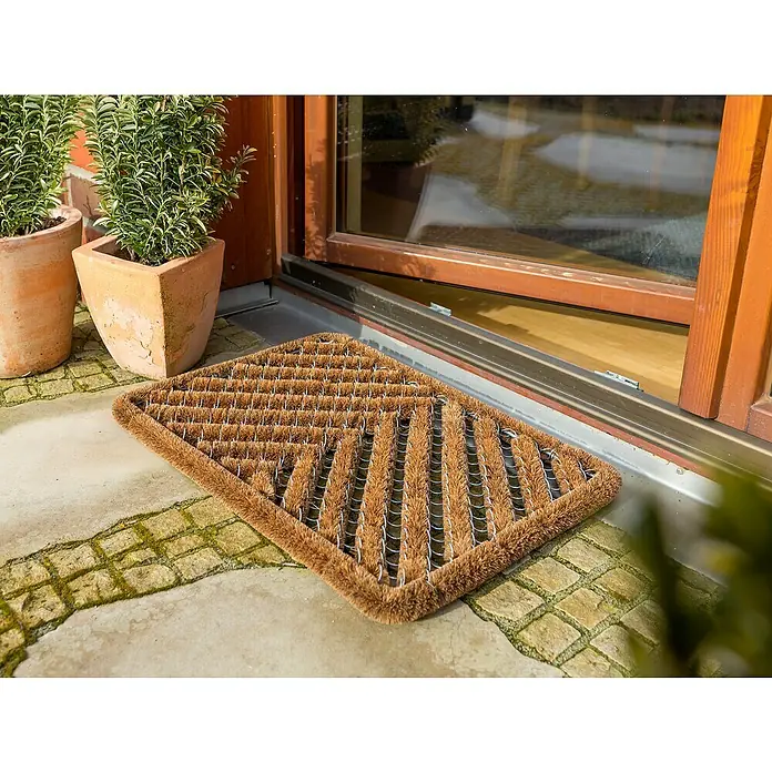 Astra Coco Fußmatte Brush - 40 x 60 cm, Kokos, Fischgrät - Eine braune Kokosfaserfußmatte liegt vor einer Haustür auf einem gepflasterten Bereich mit Pflanzen in Tontöpfen.