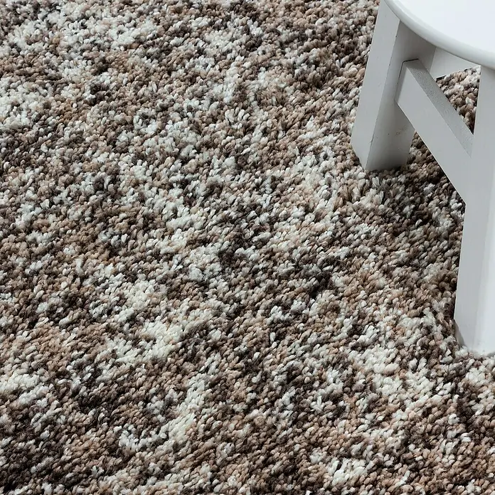 Hochflorteppich Enjoy 4500Beige, 200 x 140 cm, 100 % Polypropylen - Nahaufnahme eines mehrfarbigen Teppichs mit einem Hockerbein. Das Muster besteht aus Braun-, Beige- und Weißtönen.