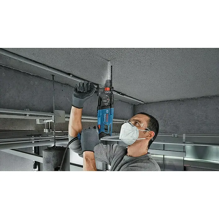 Bosch Professional Kombihammer GBH 2-21 - 720 W, 2 J, SDS-Plus-Aufnahme, Inkl. 3 x SDS-Plus Bohrer und Koffer - Ein Bauarbeiter bohrt mit einer blauen Bosch Professional Bohrmaschine ein Loch in eine Betondecke. Er trägt Schutzbrille, Maske und Handschuhe.