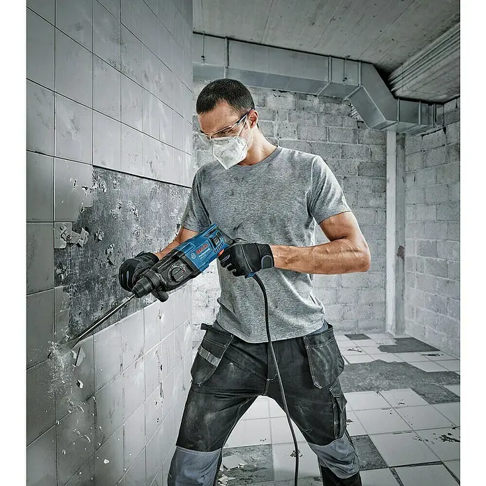 Bosch Professional Kombihammer GBH 2-21 - 720 W, 2 J, SDS-Plus-Aufnahme, Inkl. 3 x SDS-Plus Bohrer und Koffer - Ein Handwerker benutzt einen Bosch Bohrhammer, um Fliesen von einer Wand zu entfernen. Der Bosch Bohrhammer ist ein robustes Werkzeug für Bauarbeiten.
