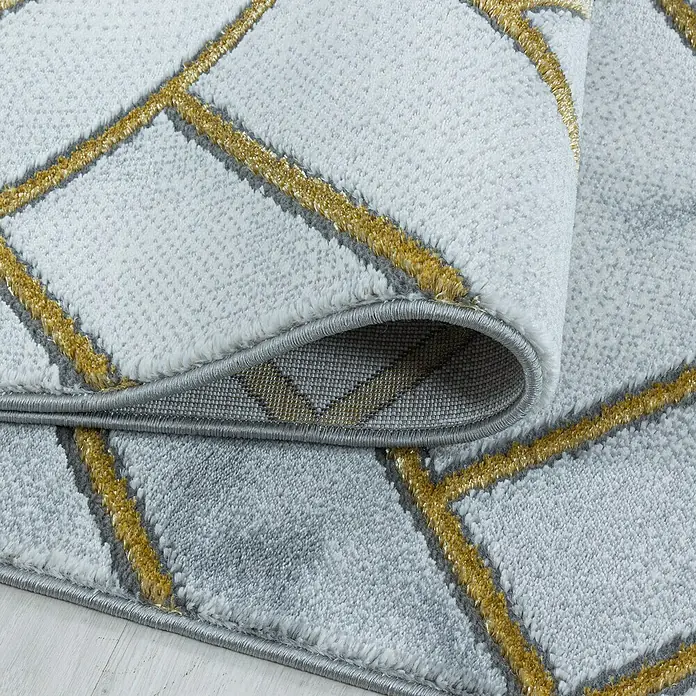 Kurzflorteppich Naxos 3813 - Gold, 200 x 140 cm, 80% Polypropylen, 20% Polyester - Nahaufnahme eines modernen Teppichs mit geometrischem Muster in Grau und Gold, teilweise aufgerollt, um die Unterseite zu zeigen.