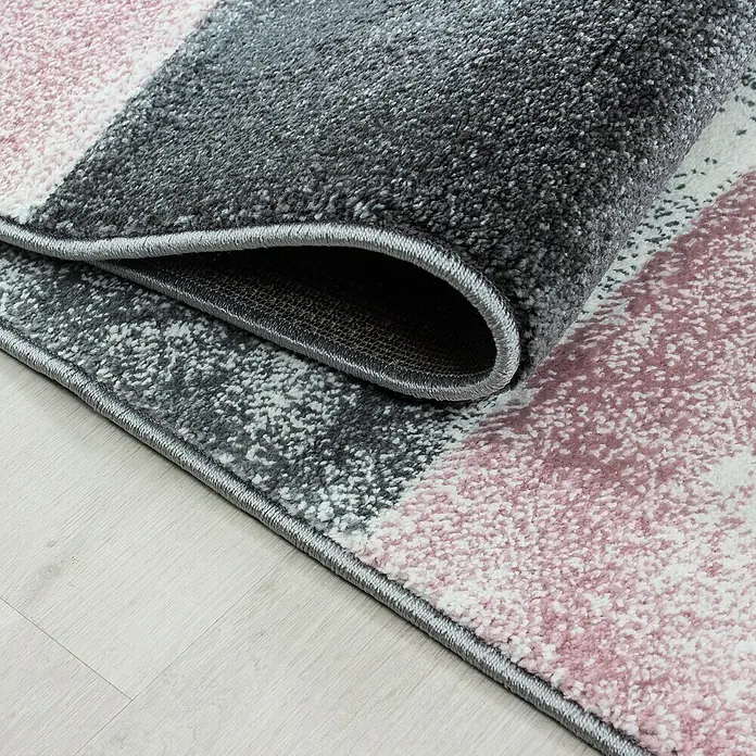 Kurzflorteppich Hawaii 1710Pink, 150 x 80 cm, 100% Polypropylen - Detailaufnahme eines modernen Teppichs mit rosa und grauen Streifen und aufgerollter Ecke, die die Textur und Verarbeitung zeigt.