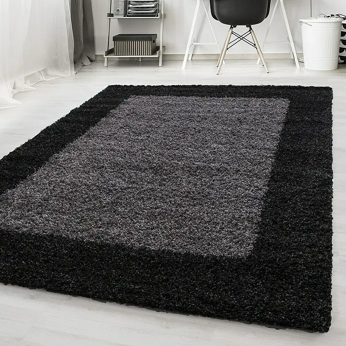 Hochflorteppich Life 1503Anthrazit, 400 x 300 cm, 100 % Polypropylen - Ein grauer Teppich mit schwarzem Rand liegt auf einem hellen Holzboden in einem Raum. Möbel sind im Hintergrund zu sehen.