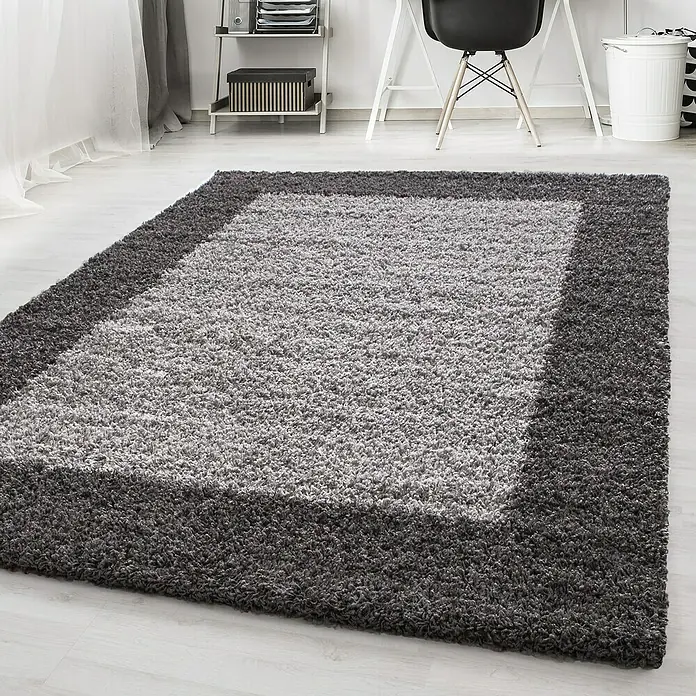 Hochflorteppich Life 1503Grau, 230 x 160 cm, 100% Polypropylen - Nahaufnahme eines grauen, rechteckigen Shaggy-Teppichs mit dunklerem Rand vor einem Schreibtisch und Regalen.