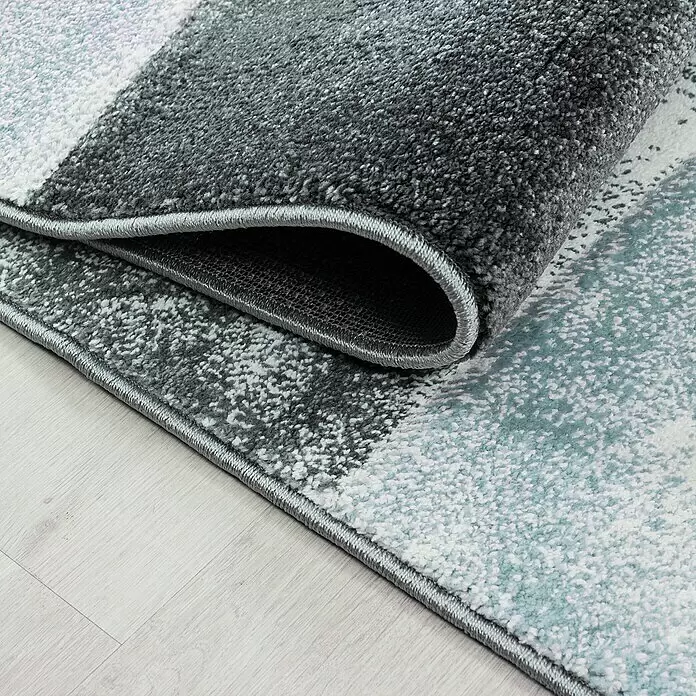 Kurzflorteppich Hawaii 1710 - Blau, 230 x 160 cm, 100 % Polypropylen - Teilweise aufgerollter Teppich mit gesprenkeltem Muster in Grau, Weiß und Hellblau auf Holzboden.
