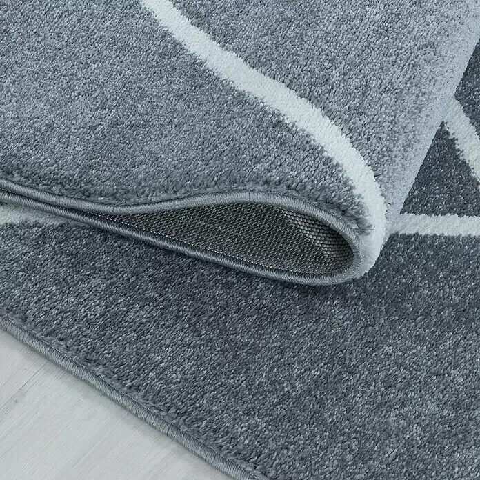 Hochflorteppich Rio 4601 - Silber, 150 x 80 cm, 100 % Polypropylen - Nahaufnahme eines grauen Teppichs mit weißen geometrischen Mustern, teilweise gerollt, um die weiche Textur und rutschfeste Rückseite zu zeigen.