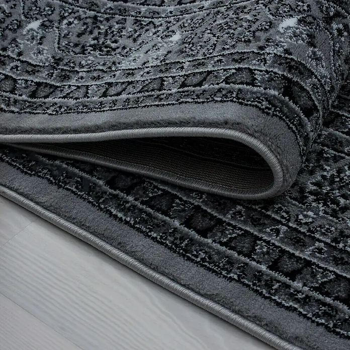 Kurzflorteppich Orient-Optik Marrakesh 0207 - Grau, 340 x 240 cm, 100% Polypropylen - Nahaufnahme eines grauen Teppichs mit gewickeltem Rand, der das Detail und die Textur des Produkts hervorhebt.