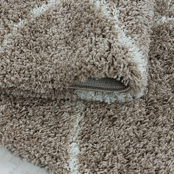Hochflorteppich Salsa 3201Beige, 200 x 140 cm, 100% Polypropylen - Nahaufnahme eines beigen Shaggy-Teppichs mit hellem Streifenmuster und aufgerollter Ecke, der die Struktur und den Flor zeigt.