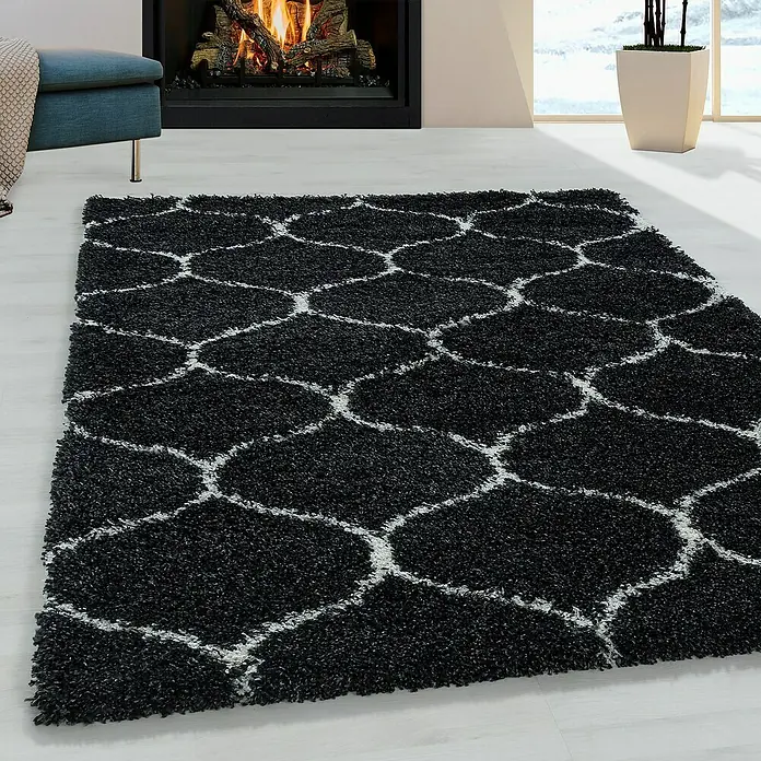Hochflorteppich Salsa 3201Anthrazit, 290 x 200 cm, 100% Polypropylen - Ein schwarzer Teppich mit einem weißen geometrischen Muster liegt vor einem Kamin und einem blauen Sessel.