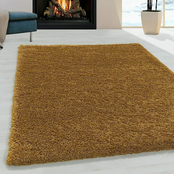 Hochflorteppich Sydney 3000 - Gold, 200 x 100 cm, 100 % Polypropylen - Nahaufnahme eines zotteligen, ockerfarbenen Teppichs, der auf einem hellen Holzboden vor einem Kamin und einer Fensterbank liegt.