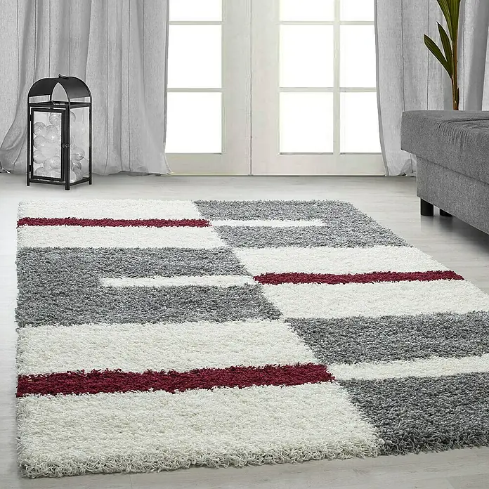 Hochflorteppich Gala 2505Rot, 110 x 60 cm, 100 % Polypropylen - Ein flauschiger Teppich mit geometrischen Mustern in Grau, Weiß und Burgunderrot. Der Teppich liegt in einem hellen Raum mit Dekorationen.