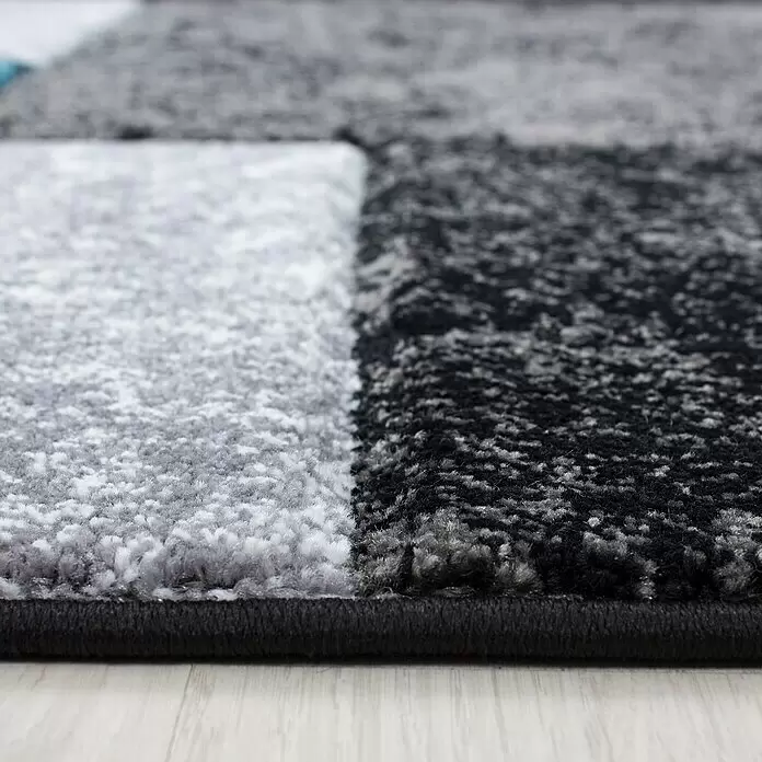 Kurzflorteppich Hawaii 1330 - Türkis, 340 x 240 cm, 100 % Polypropylen - Nahaufnahme eines strukturierten grau-schwarz-weißen Teppichs mit geometrischem Muster und dunkler Bordüre auf hellem Holzboden.