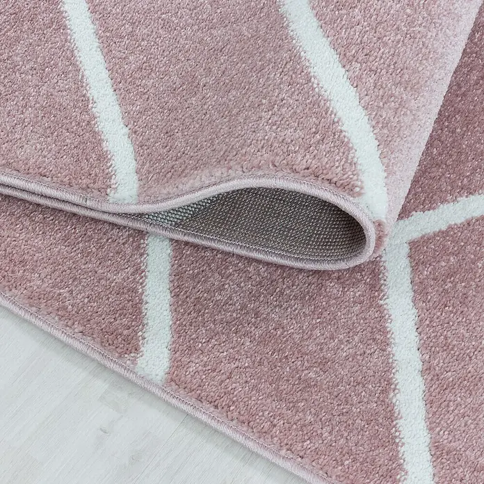Hochflorteppich Rio 4602 - Rose, 200 x 140 cm, 100% Polypropylen - Detailaufnahme eines rosa Teppichs mit geometrischen weißen Streifen und aufgerollter Kante, der seine Textur und Verarbeitung zeigt.