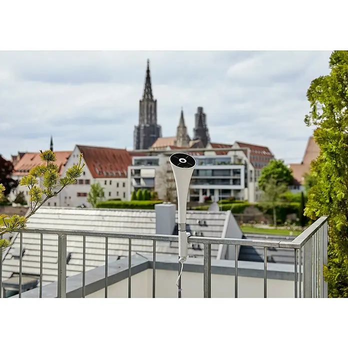 Gardena ClickUp! Solarleuchte - Ø x H: 11 x 30,7 cm - Das Bild zeigt einen weißen tragbaren Lautsprecher, der an einem Balkongeländer befestigt ist, mit einer Stadtlandschaft im Hintergrund.