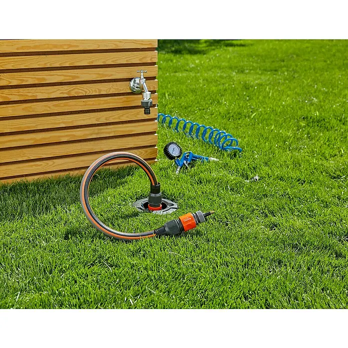 Gardena Sprinklersystem Wassersteckdose - ¾″ - Ein orangefarbener Gartenschlauch mit schwarzem Adapter liegt auf dem Rasen vor einem Wasserhahn und einem blauen Luftschlauch.