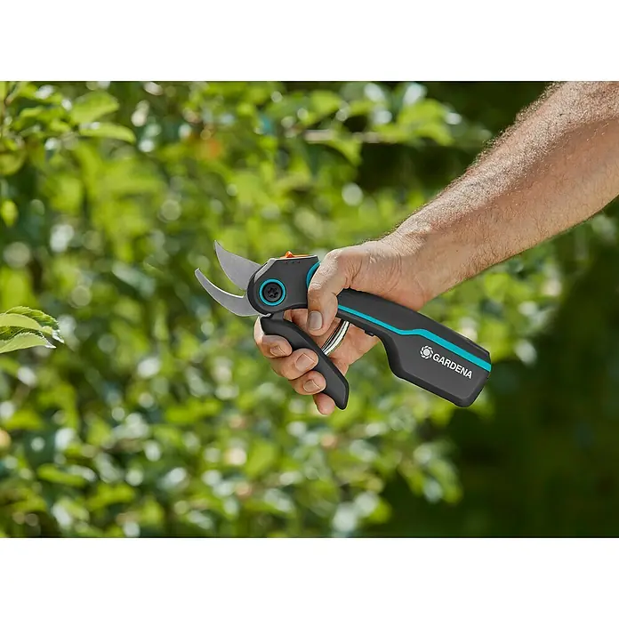 Gardena Akku-Strauchschere AssistCut - L x B x H: 270 x 70 x 30 mm, Max. Aststärke: 25 mm - Eine Hand hält eine Gardena Gartenschere vor einem grünen Hintergrund. Die Schere ist schwarz mit türkisfarbenen Akzenten.