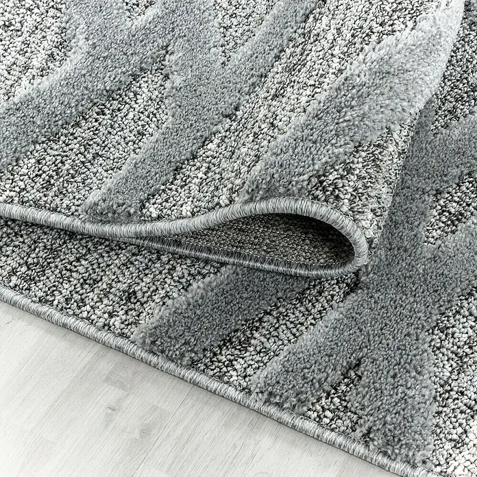 Hochflorteppich Pisa 4706Grau, 340 x 240 cm, 100% Polypropylen - Detailaufnahme eines grauen Teppichs mit abstraktem Muster und umgeschlagenem Rand. Der Teppich liegt auf einem hellen Holzboden.