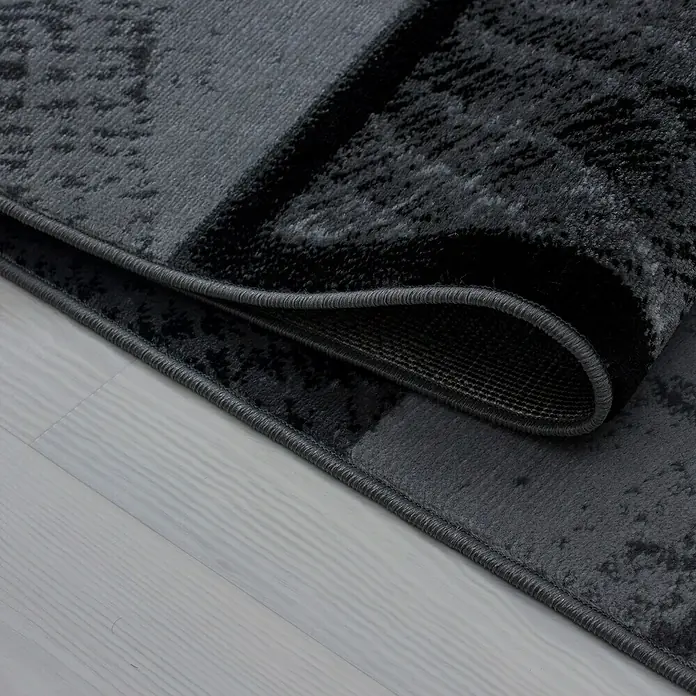 Kurzflorteppich Plus - Schwarz/Anthrazit, 370 x 280 cm, 100 % Polypropylen - Nahaufnahme eines modernen, grauen Teppichs mit abstraktem Muster und umgeschlagener Ecke, der die Textur und Verarbeitung zeigt.