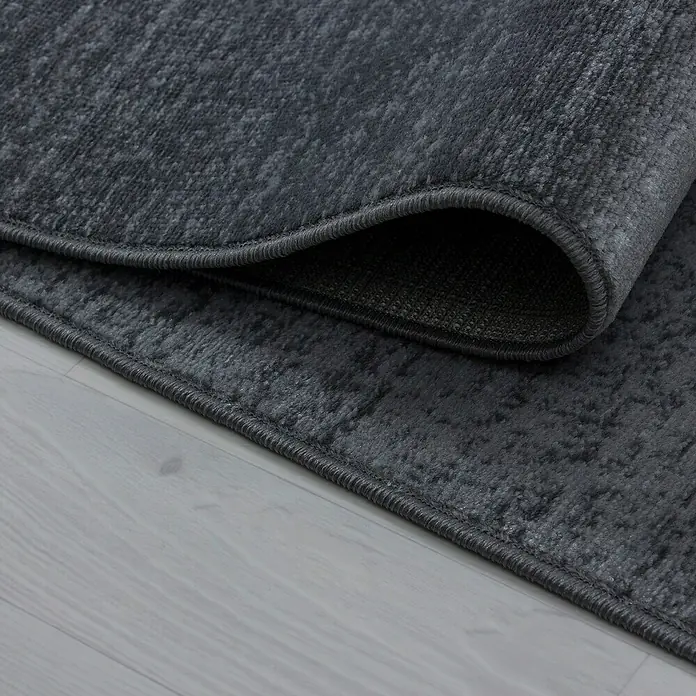 Kurzflorteppich Plus 8002 - Grau, 230 x 160 cm, 100% Polypropylen - Detailaufnahme eines grauen Teppichs mit gewellter Kante, der auf einem hellen Holzboden liegt.