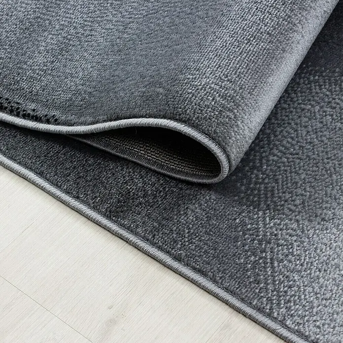 Kurzflorteppich Plus 8008 - Schwarz, 370 x 280 cm, 100 % Polypropylen - Nahaufnahme eines grauen Teppichs mit aufgerolltem Rand, der Textur und Detailgenauigkeit zeigt.