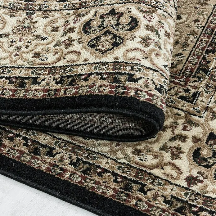 Kurzflorteppich Kashmir 2608Schwarz, 230 x 160 cm, 100% Polypropylen - Detailaufnahme eines eleganten Teppichs mit floralem Muster und schwarzer Bordüre, teilweise aufgerollt, um die Textur zu zeigen.