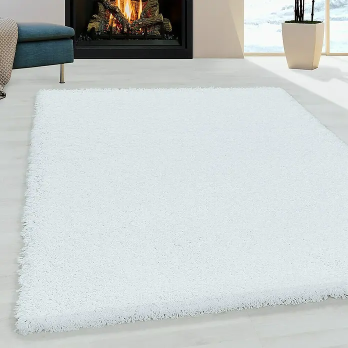 Hochflorteppich Sydney 3000Weiß, 170 x 120 cm, 100% Polypropylen - Ein weißer, rechteckiger Hochflorteppich liegt auf einem hellen Holzboden vor einem Kamin, der ein gemütliches Ambiente schafft.