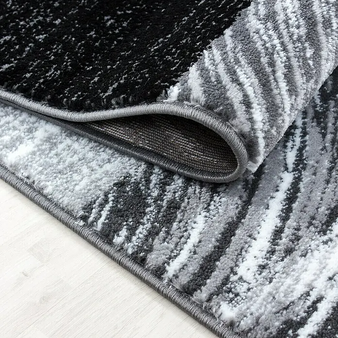 Kurzflorteppich Parma 9220Schwarz, 150 x 80 cm, 100% Polypropylen - Nahaufnahme eines modernen Teppichs mit abstraktem Muster in Schwarz, Grau und Weiß, teilweise aufgerollt, um die Textur zu zeigen.