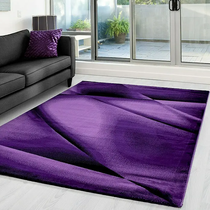 Kurzflorteppich Miami 6590 - Lila, 290 x 200 cm, 100% Polypropylen - Ein lila Teppich mit einem abstrakten Muster liegt in einem Wohnzimmer, neben einem grauen Sofa und einem großen Fenster.