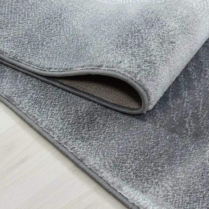 Kurzflorteppich Parma 9310 - Schwarz, 300 x 80 cm, 100% Polypropylen - Nahaufnahme eines grauen Teppichs mit gewalztem Rand und brauner Unterseite, der auf einem hellen Holzboden liegt.