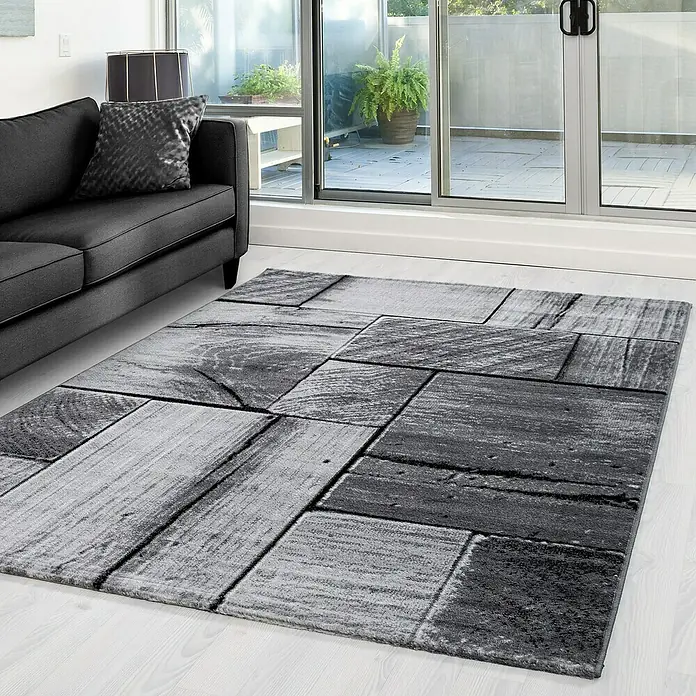 Kurzflorteppich Parma 9260 - Schwarz, 300 x 80 cm, 100% Polypropylen - Ein grauer Teppich mit einem modernen Holzblock-Design liegt in einem hellen Raum mit einem Sofa und großen Fenstern.