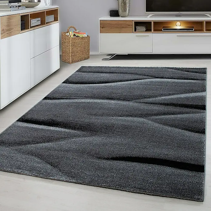Kurzflorteppich Lucca 1840Schwarz, 170 x 120 cm, 100 % Polypropylen - Ein grauer Teppich mit gewellten Linien liegt in einem modernen Wohnzimmer, ergänzt die Einrichtung mit weißem Sideboard und TV-Möbel.