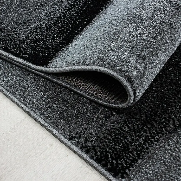 Kurzflorteppich Lucca 1840Schwarz, 170 x 120 cm, 100 % Polypropylen - Nahaufnahme eines grau-schwarzen Teppichs mit gerolltem Rand, der seine Textur und Verarbeitung zeigt.