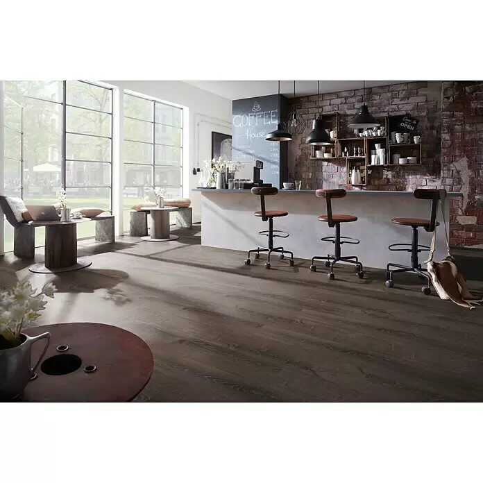 Star Clic Vinylboden More+ - Smoky Oak Grey, Landhausdiele, 1.210 x 220 x 5 mm - Dunkler Bodenbelag in Holzoptik in einem modernen Café im Industriestil mit großen Fenstern und Ziegelwänden.