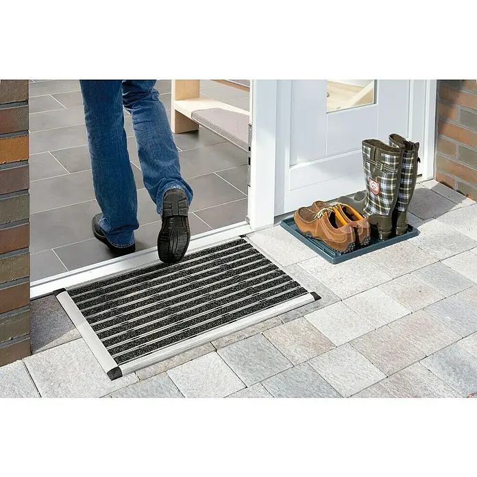 Astra Fußmatte Excellence Mat - Aluminium, Anthrazit/Grau, 40 x 60 cm - Ein Fuß betritt einen Eingangsmattenabstreifer vor einer Haustür, mit Schuhen und Stiefeln auf einer Ablage daneben.