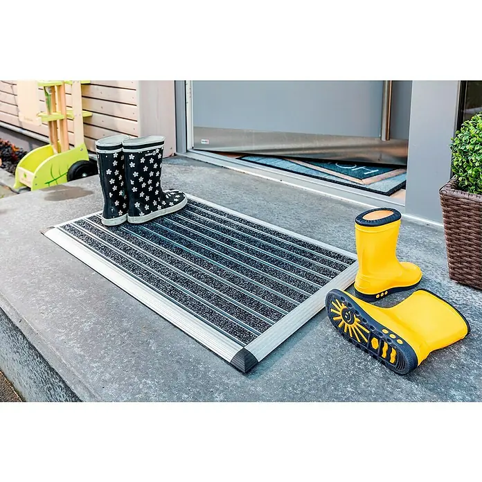 Astra Fußmatte Excellence Mat - Aluminium, Anthrazit/Grau, 40 x 60 cm - Eine Fußmatte mit Aluminiumrahmen und zwei Paar Gummistiefeln, eines schwarz mit Blumenmuster und eines gelb, vor einer Haustür.