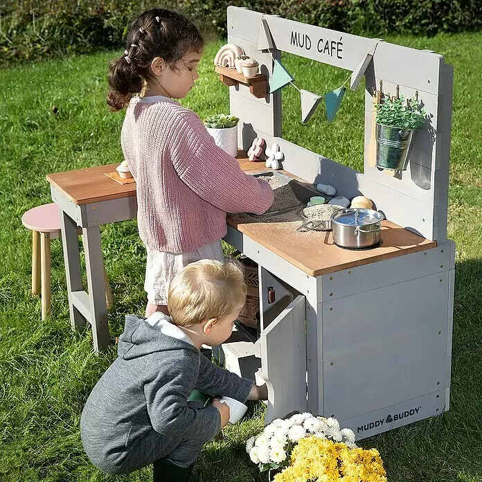 Muddy Buddy Matschküche Mud Café - 110 x 71,5 x 100 cm, Holz, Warmgrau - Zwei Kinder spielen draußen mit einer grauen Holzküche "Mud Café".