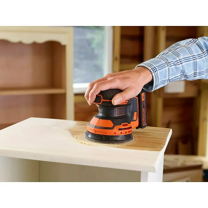 Black+Decker Akku-Exzenterschleifer BDCROS18-QW - 18 V, Li-Ionen, 1,5 Ah, 1 Akku, 125 mm - Nahaufnahme eines Black+Decker Akku-Schleifers, der auf einer Holzoberfläche verwendet wird. Das Werkzeug ist orange und schwarz und wird von einer Hand gehalten.