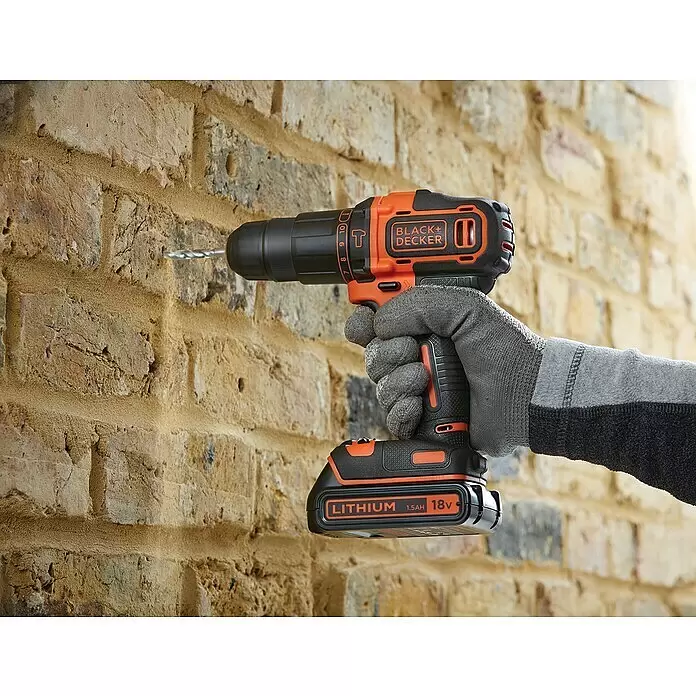 Black+Decker Taladro atornillador percutor de batería BDCHD18KB - 18 V, 2 baterías, 1,5 Ah, 40 Nm - Mano enguantada sosteniendo un taladro inalámbrico Black+Decker naranja y negro con batería de litio de 18V, perforando una pared de ladrillos.