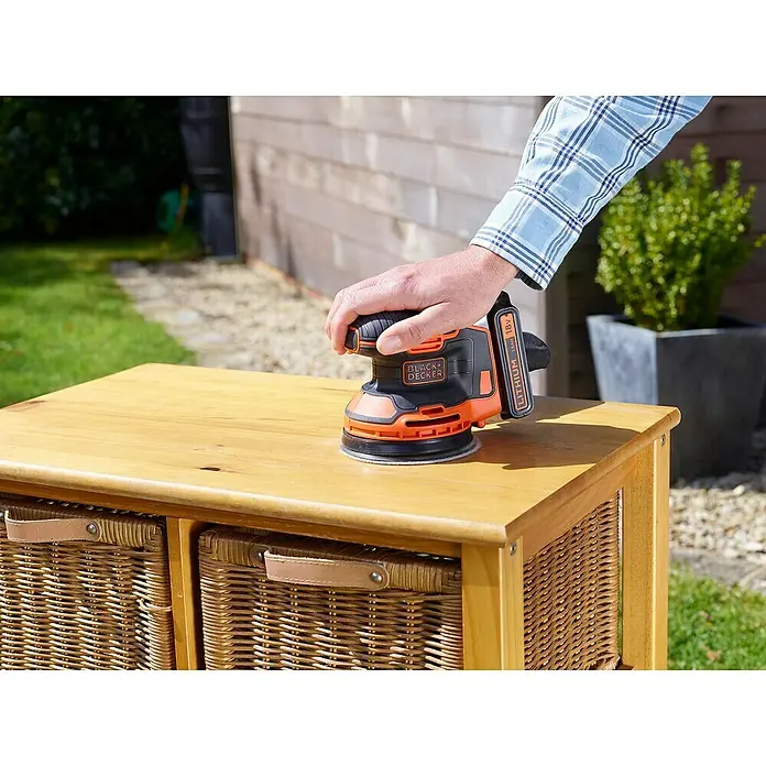 Black+Decker Akku-Exzenterschleifer BDCROS18-QW - 18 V, Li-Ionen, 1,5 Ah, 1 Akku, 125 mm - Nahaufnahme eines Black+Decker-Schleifers, der zum Glätten einer Holzoberfläche auf einem Holztisch mit geflochtenen Körben verwendet wird.