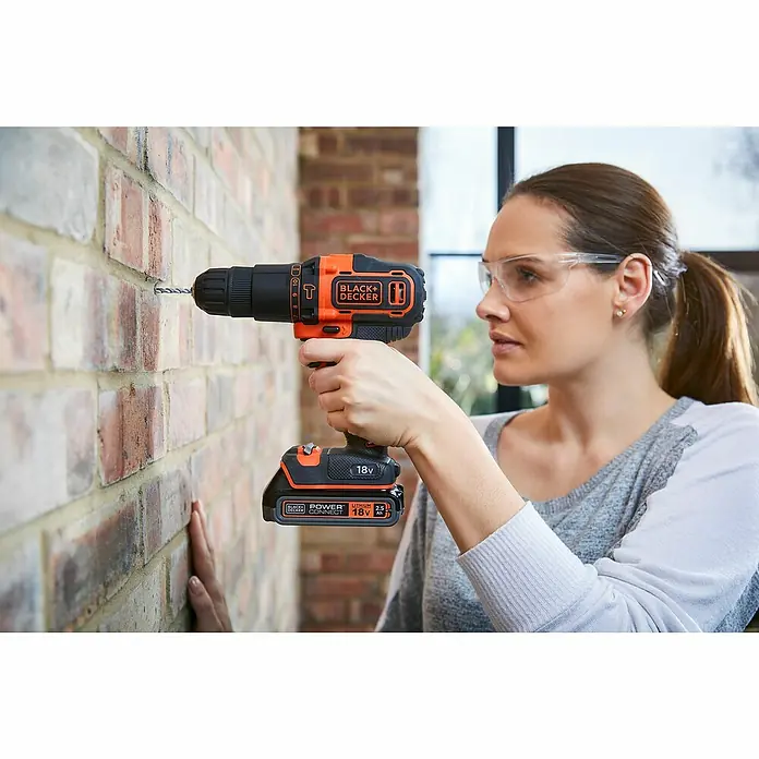 Black+Decker Taladro atornillador percutor de batería BDCHD18KB - 18 V, 2 baterías, 1,5 Ah, 40 Nm - Mujer perfora una pared de ladrillos con un taladro inalámbrico Black+Decker, con gafas de seguridad.