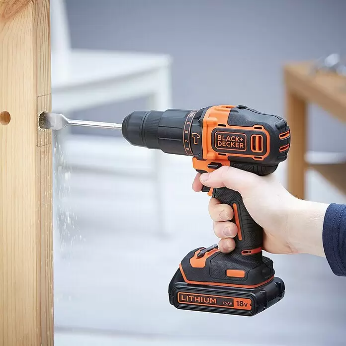 Black+Decker Taladro atornillador percutor de batería BDCHD18KB - 18 V, 2 baterías, 1,5 Ah, 40 Nm - Una persona utiliza un taladro inalámbrico Black+Decker naranja y negro con una broca de pala para perforar un agujero en una viga de madera clara. Vuela aserrín.