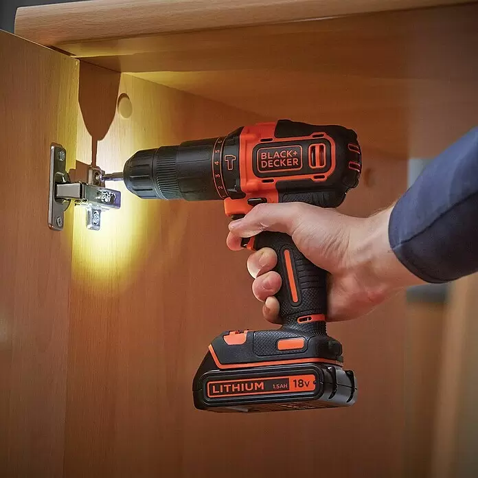Black+Decker Taladro atornillador percutor de batería BDCHD18KB - 18 V, 2 baterías, 1,5 Ah, 40 Nm - Taladro inalámbrico BLACK+DECKER negro y naranja en mano, atornillando una bisagra en un mueble de madera. Su luz ilumina el área de trabajo.