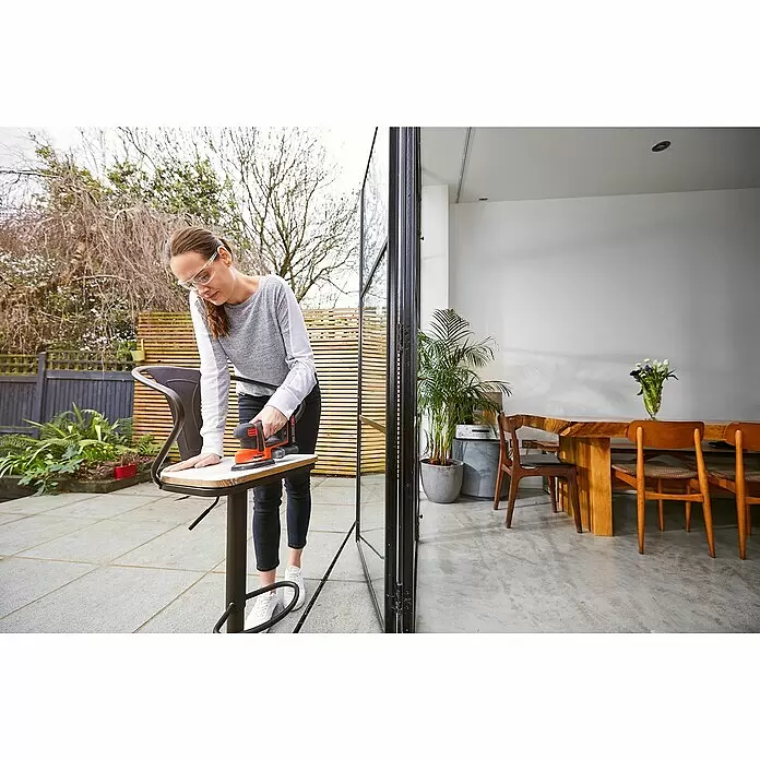 Black+Decker 18V PowerConnect Akku-Deltaschleifer Mouse® BDCDS18N-XJ - 18 V, Li-Ionen, Ohne Akku, 24.000 U/min - Eine Frau verwendet einen orange-schwarzen Schleifer, um einen Holzschemel im Freien zu glätten. Sie trägt eine Schutzbrille.