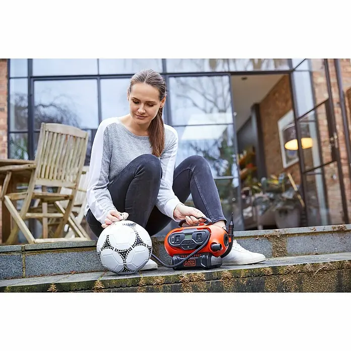 Black+Decker Akku-Kompressor BDCINF18N-QS - 18 V, Ohne Akku, 11 bar - Eine Frau sitzt draußen und pumpt einen Fußball mit einem tragbaren, orangefarbenen Black+Decker Luftkompressor auf.